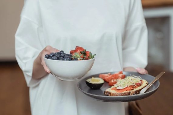Person holding a bowl of fresh berries and a plate with avocado toast, displaying a healthy breakfast spread.